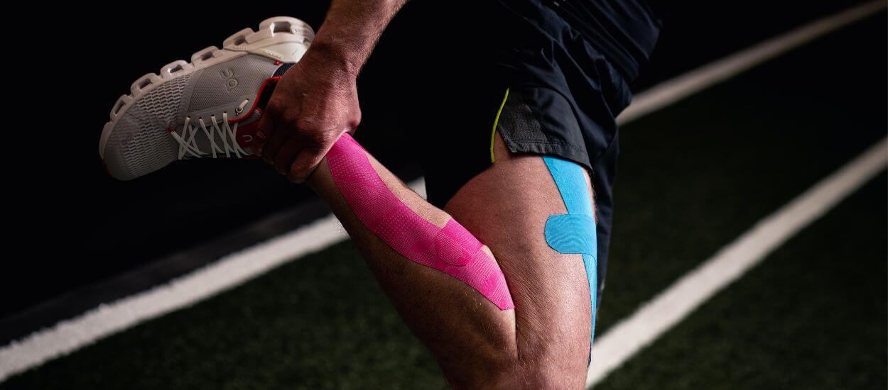 A close-up image of an athlete's upper leg, showing pink and blue kinesiology tape applied to the skin. The athlete is wearing black shorts with yellow trim, and the background is a blurred sports field.